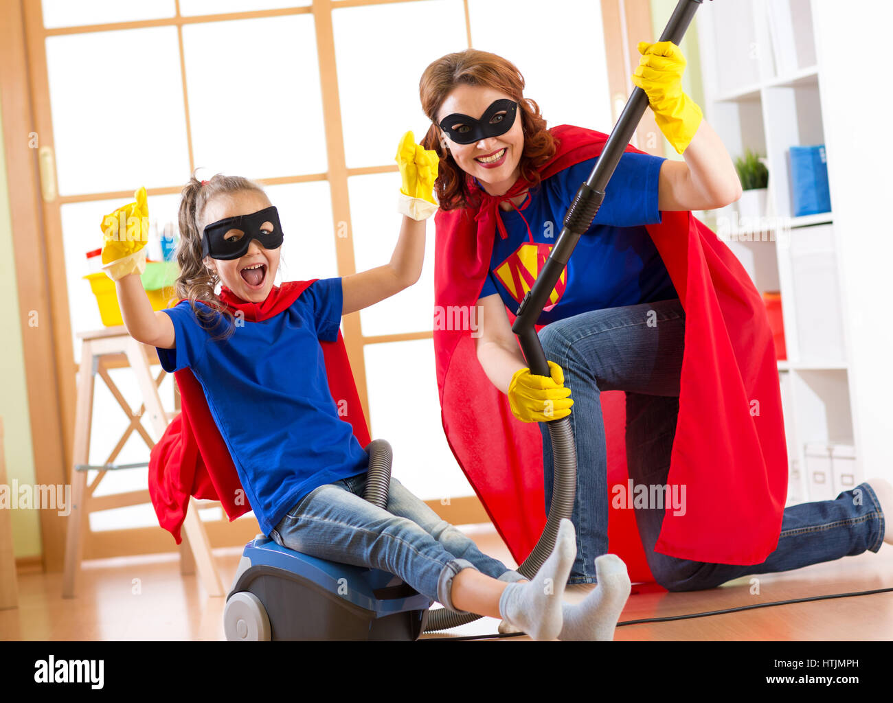 Kind und Mutter verkleidet als Superhelden mit Staubsauger im Zimmer. Familie von mittlerem Alter Frau und Tochter viel Spaß während der Reinigung des Bodens. Stockfoto