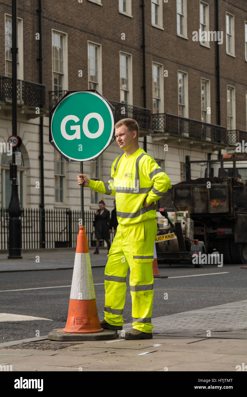Ein gut sichtbarer Straßenarbeiter mit einem grünen „GO“-Schild, der den Verkehr in London leitet Stockfoto