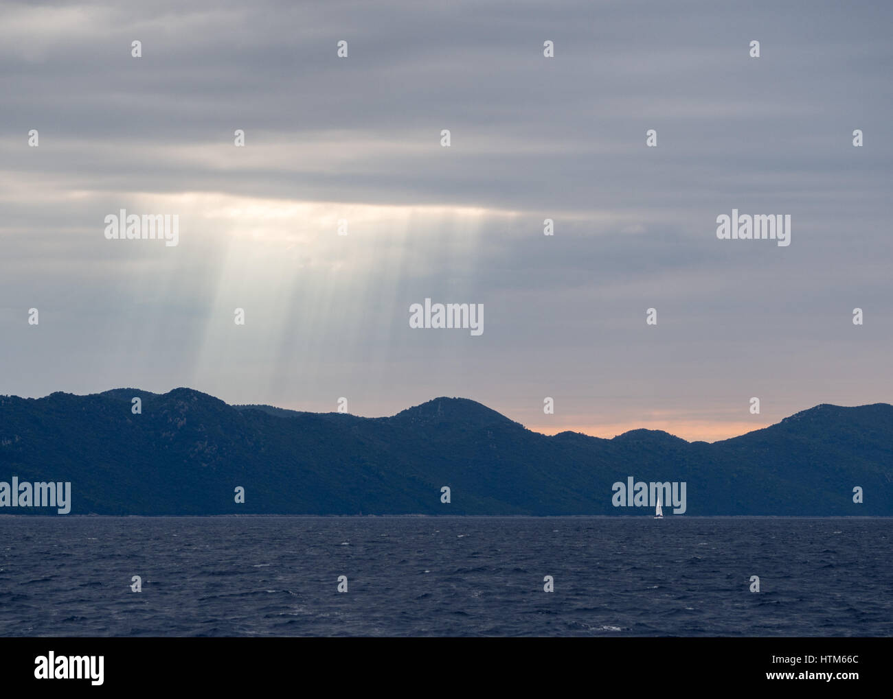 Sonnenstrahlen durch stürmische dunkle Wolken über eine Silhouette von Hügeln und Bergen Berg bei Sonnenuntergang mit Yacht am Meer Ozean, Kroatien Stockfoto
