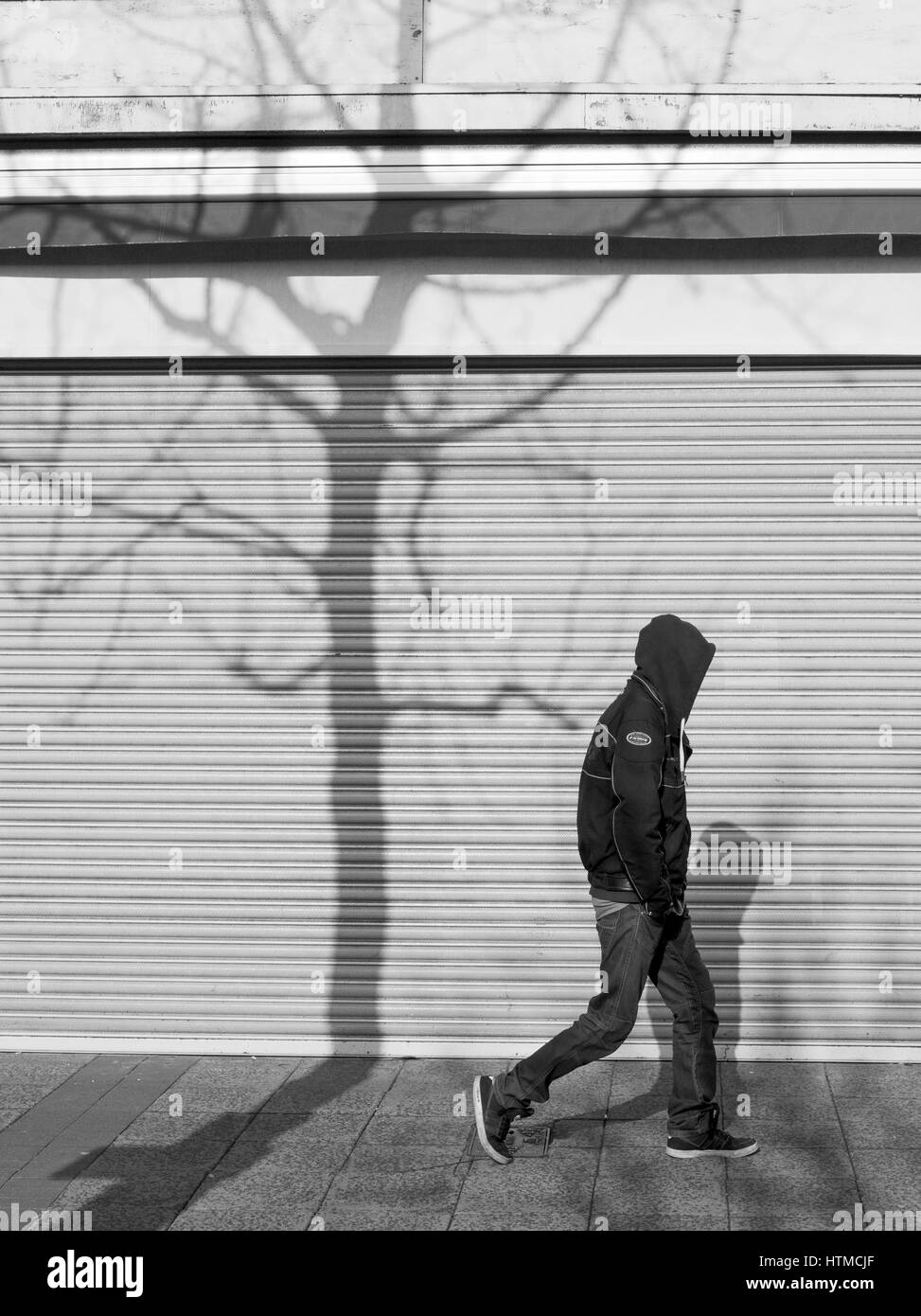 Jungen Mann mit einem Hoodie vorbeigehen Closed Shop Fensterläden mit dem Schatten eines Baumes.  Straßenfotografie in Plymouth Devon England UK Stockfoto