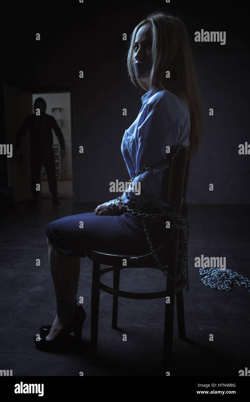 Was ist los. Mit Ketten gefesselt Angst machtlos ängstliche Frau auf einem Stuhl sitzend während ihrem Entführer beim Betreten des Zimmers Stockfoto