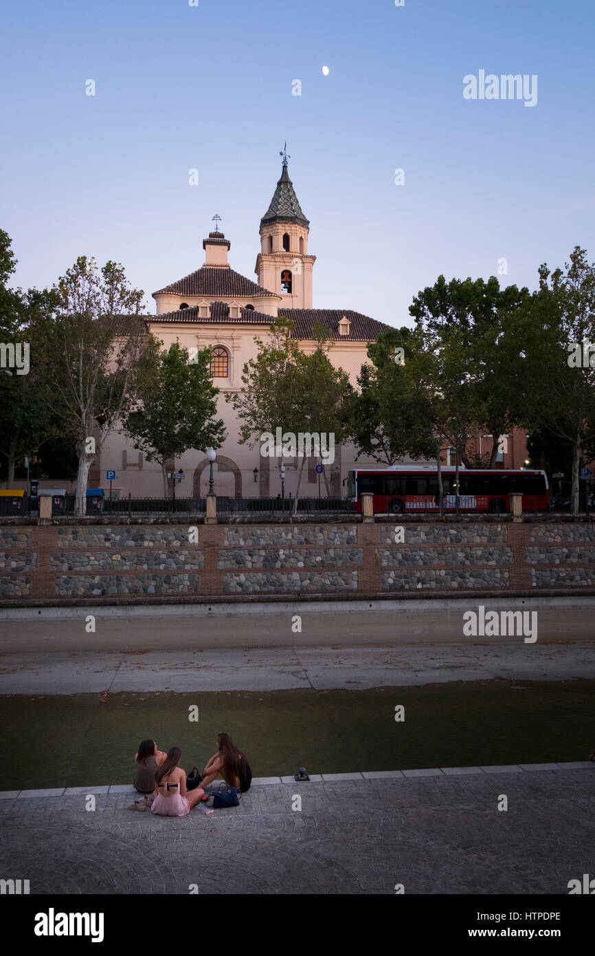 Drei junge Mädchen zurück zu Kamera sitzen durch den Fluss Genil gegenüber die Hermandad de Los Escolapios wie Dämmerung bei klarem Himmel der Mond zeigt Stockfoto