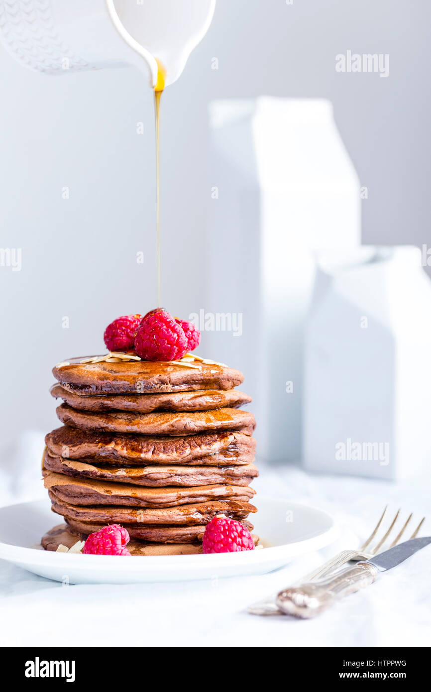 Stapel von hausgemachte amerikanische eifreie Schokolade Pfannkuchen mit Ahornsirup fließt aus Glas, serviert auf weißen Teller zusammen mit Himbeeren. Stockfoto