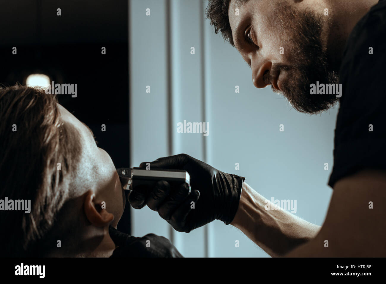 BARBERSHOP-THEMA. BÄRTIGE BARBER IN SCHWARZEN GUMMIHANDSCHUHEN IST DEN BART DES AUFTRAGGEBERS JUNGE HÜBSCHE TRIMMEN. ER NUTZT EINEN HAARSCHNEIDER Stockfoto