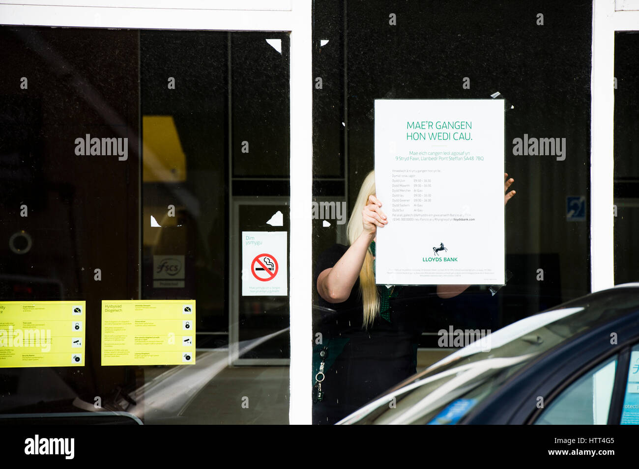 Lloyds Bank schließt seine Niederlassung in einem kleinen walisischen Stadt: 9. März 2017 - Angestellter Frau erträgt walisische Sprache Informationen Plakat im Fenster des Ortsverbandes der Lloyds Bank, am Nachmittag geschlossen für das gute, in der kleinen ländlichen Tregaron Dorf Ceredigion Wales UK Stockfoto