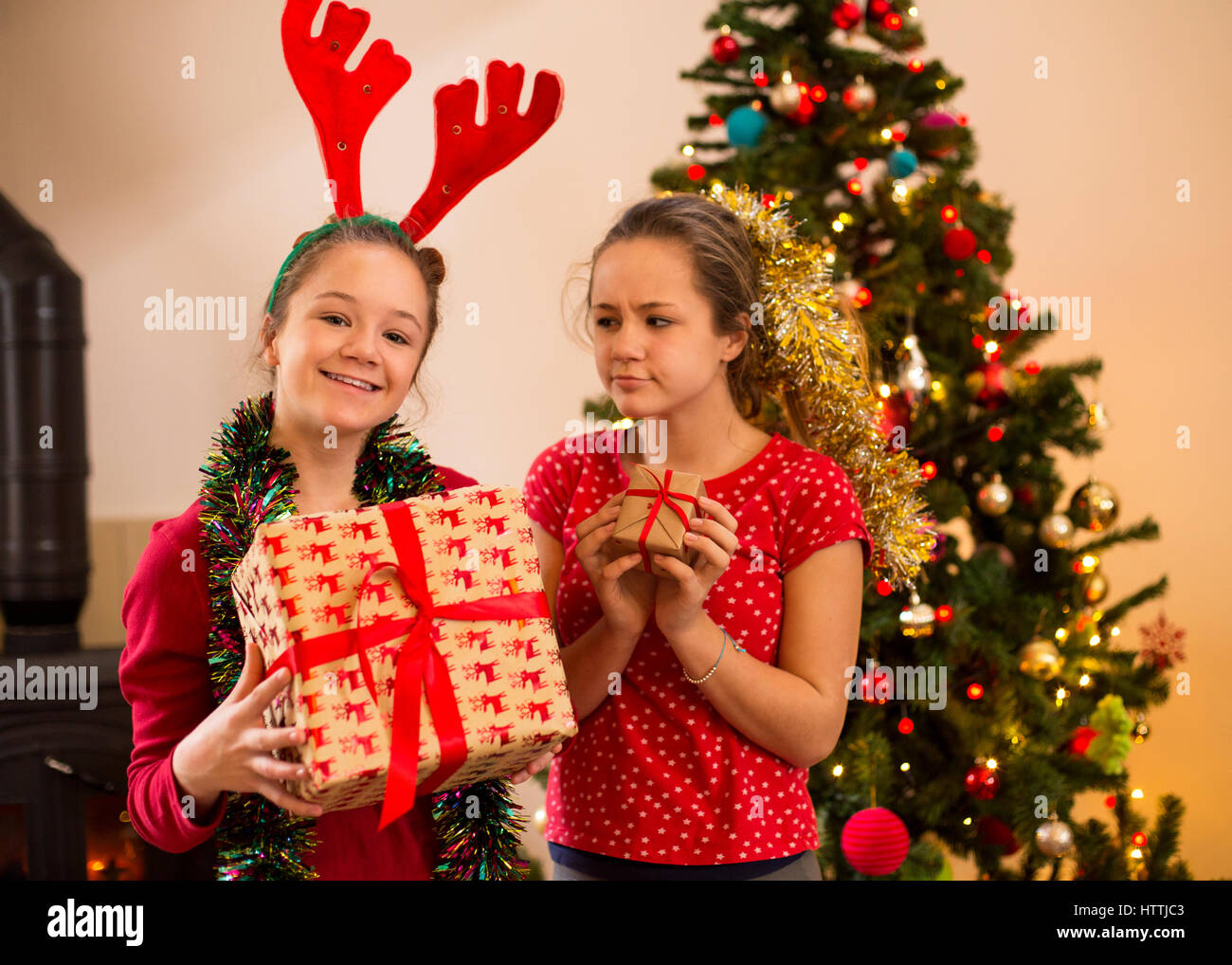 Zwei Schwestern posieren zu Weihnachten. Man hält ein großes Geschenk und das andere schaut ihr mit einem eifersüchtigen Gesicht als sie ein kleines Geschenk hält. Stockfoto
