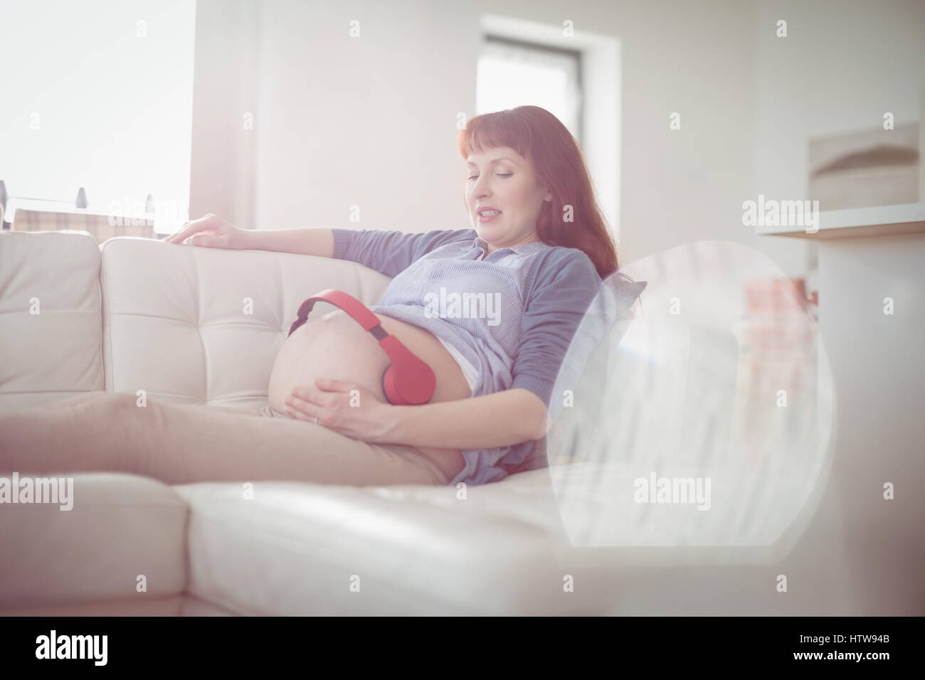 Schwangere Frau mit Kopfhörern auf den Bauch entspannen Sie sich auf sofa Stockfoto