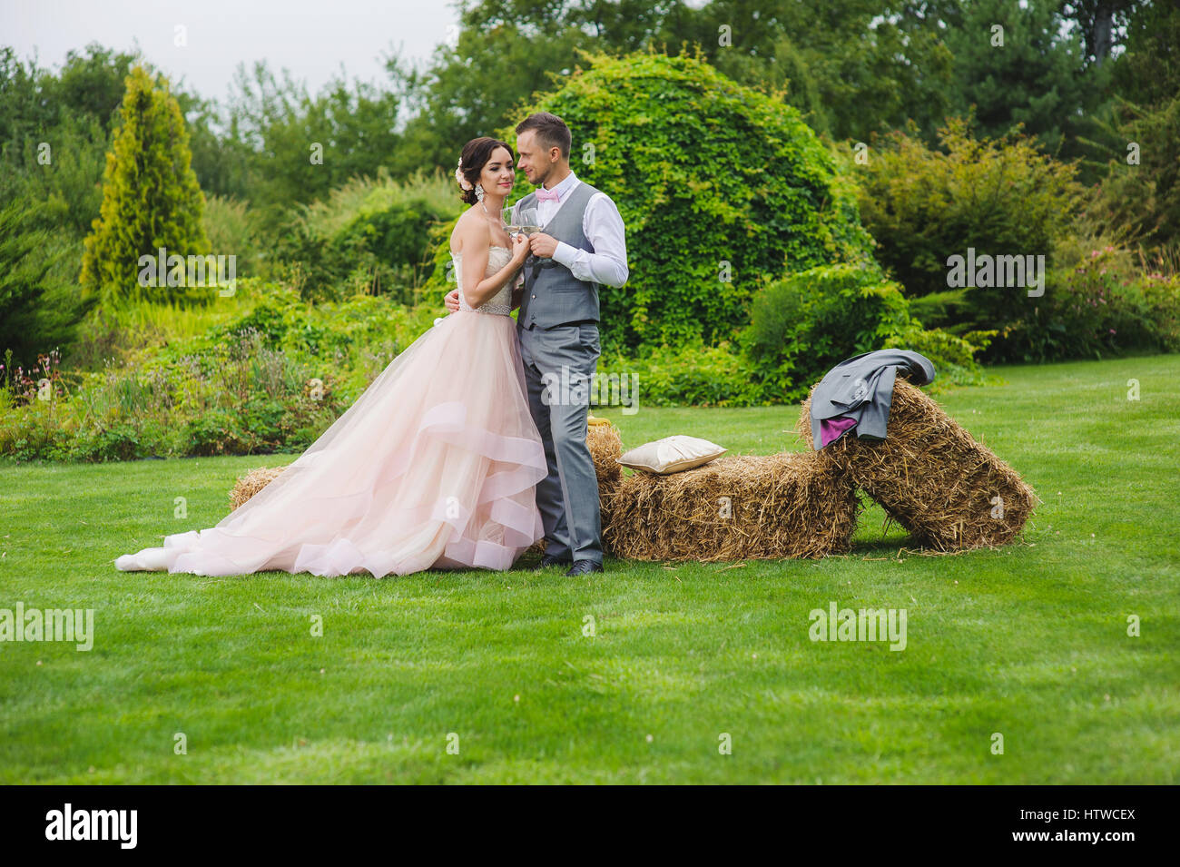 Klirrende Gläser. Braut und Bräutigam machen Toast im Freien. Grass, Ballen Heu Bäume und Büsche im Hintergrund. Stockfoto