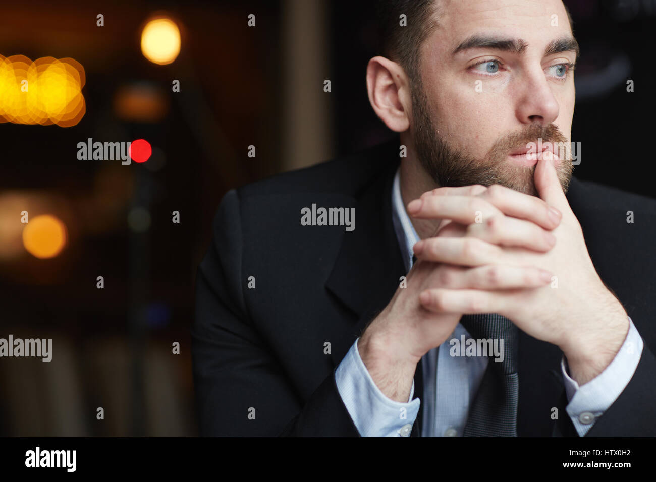 Porträt des modernen bärtigen Geschäftsmann aussehende Weg nachdenken über etwas ruhen Kopf auf gefalteten Händen vor schwarzem Hintergrund Stockfoto