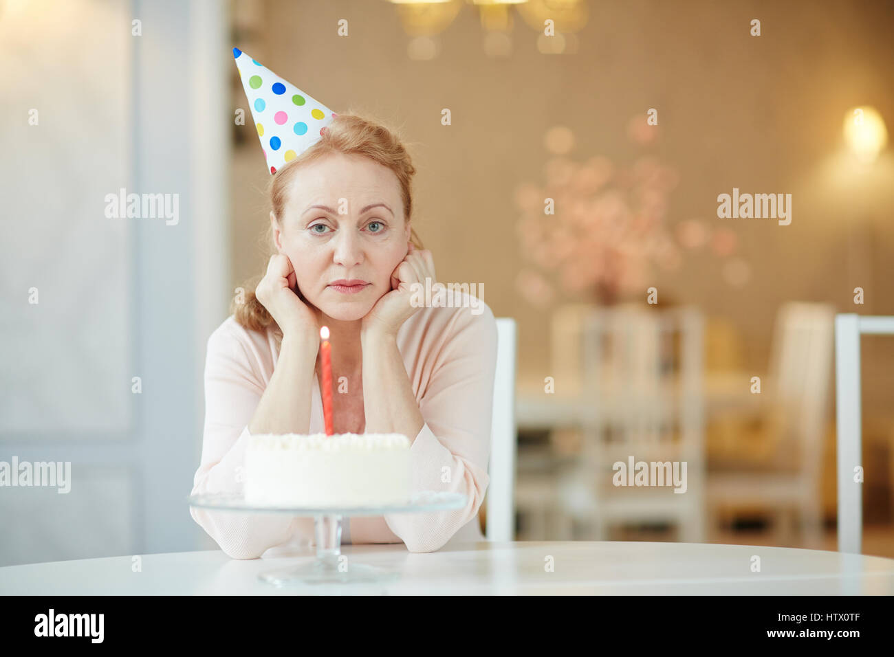 Porträt von traurig einsam Reife Frau sitzt alleine am Geburtstagstisch mit Kuchen, Party Hut und Blick in die Kamera Stockfoto