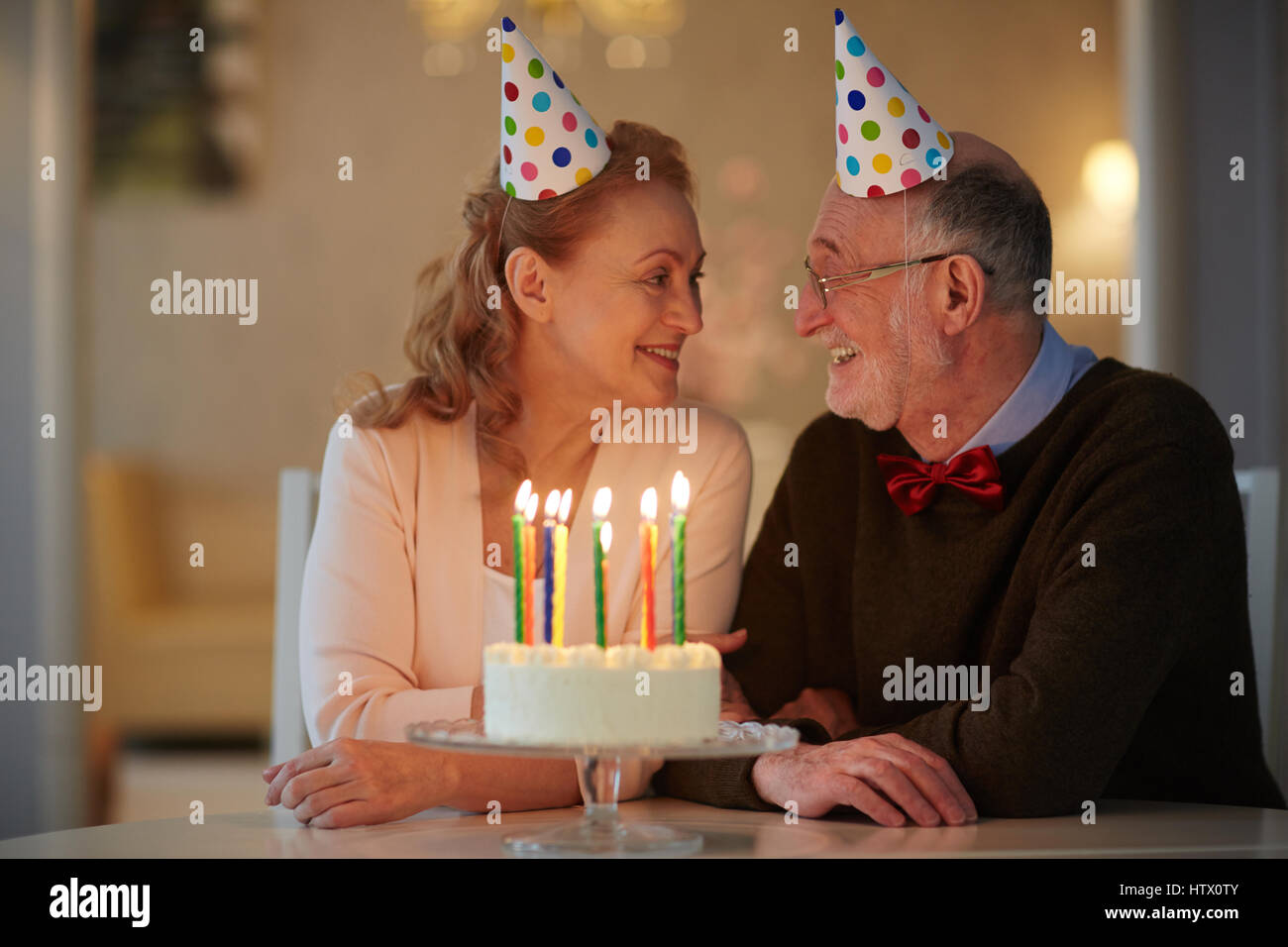 Porträt von senior Liebespaar feiert Geburtstag zusammen sitzen am Tisch mit Kuchen und tragen Partyhüte suchen Sie zärtlich in die Augen Stockfoto