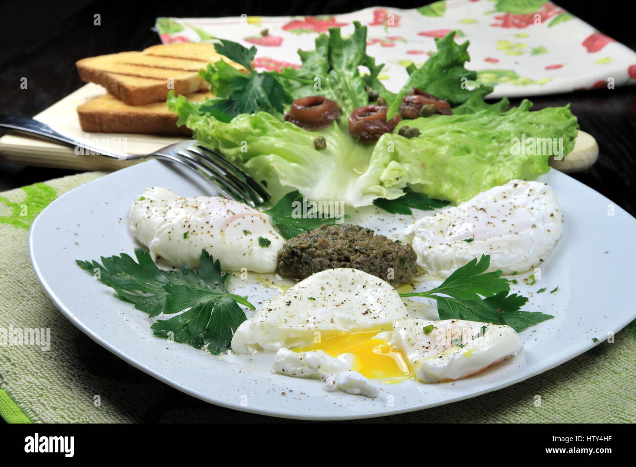 Drei pochierten Eiern auf Kapern, Petersilie und Sardellen-Sauce serviert mit Gabel in einer weißen Schale auf bunte Küchentücher - laufende gelbe Dotter auf vorderen Ei Stockfoto