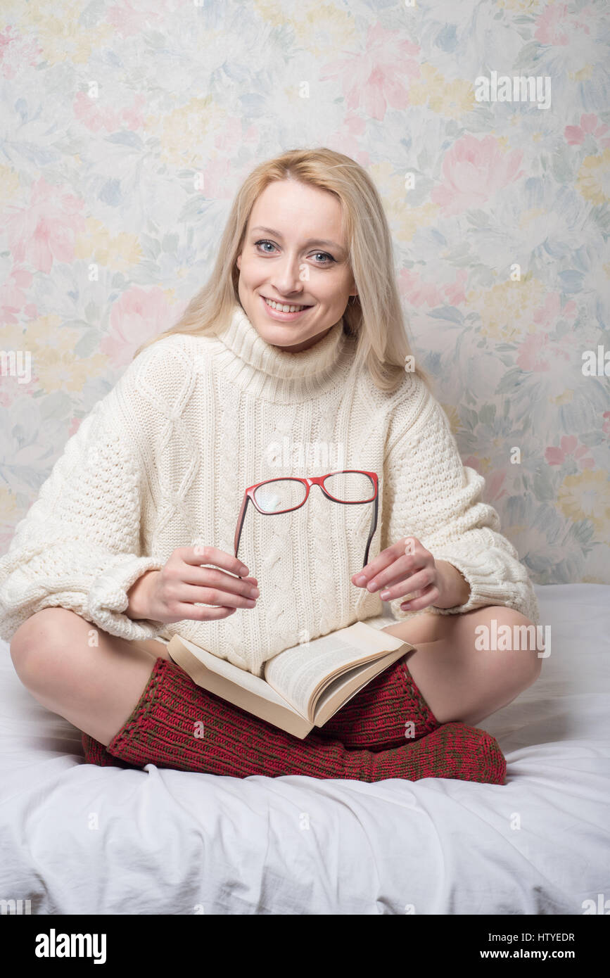 Frau sitzt im Schneidersitz auf Bett mit Buch halten Lesebrille Stockfoto