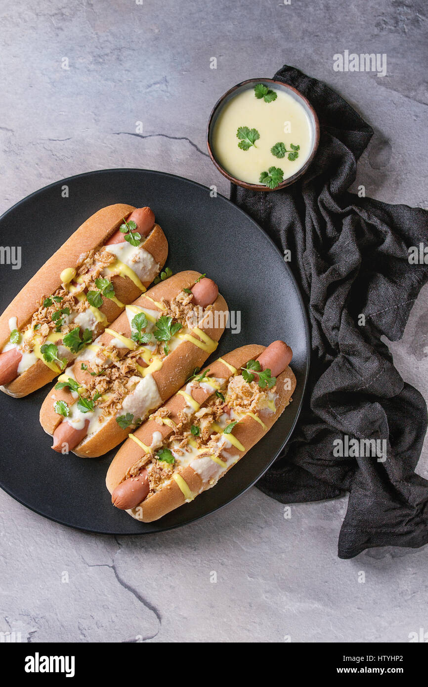 Hot Dogs mit Wurst, gebratenen Zwiebeln, Koriander, Käse-Sauce und Senf, serviert auf schwarze Keramikplatte mit Textil über graue Textur Zeitmessung Stockfoto
