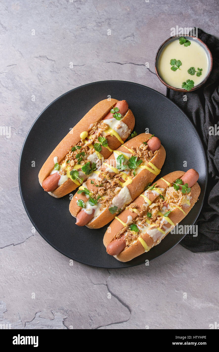 Hot Dogs mit Wurst, gebratenen Zwiebeln, Koriander, Käse-Sauce und Senf, serviert auf schwarze Keramikplatte mit Textil über graue Textur Zeitmessung Stockfoto