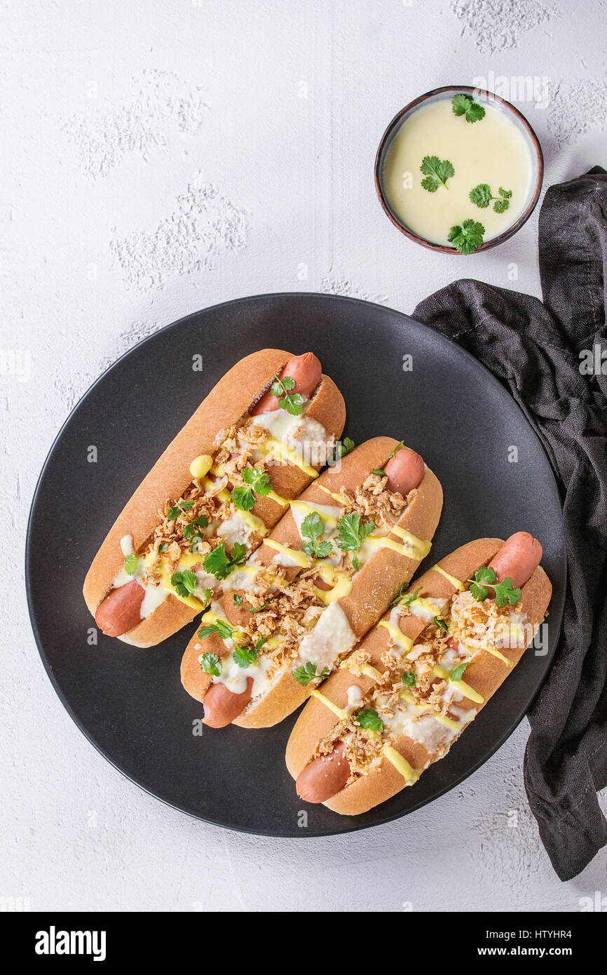 Hot Dogs mit Wurst, gebratenen Zwiebeln, Koriander, Käse-Sauce und Senf, serviert auf schwarze Keramikplatte mit Textil über weißem Beton Textur Stockfoto