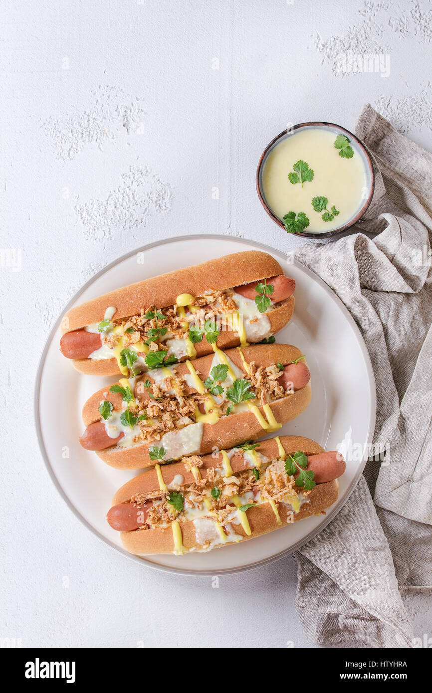 Hot Dogs mit Wurst, gebratenen Zwiebeln, Koriander, Käse-Sauce und Senf, serviert auf weißen Keramikplatte mit Textil über weißem Beton Textur Stockfoto