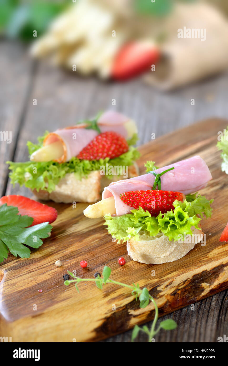 Köstliche Häppchen mit Schinken Brötchen und weißer Spargel mit frischen Erdbeeren auf knusprigem Baguette mit Blätter Lollo bionda Stockfoto