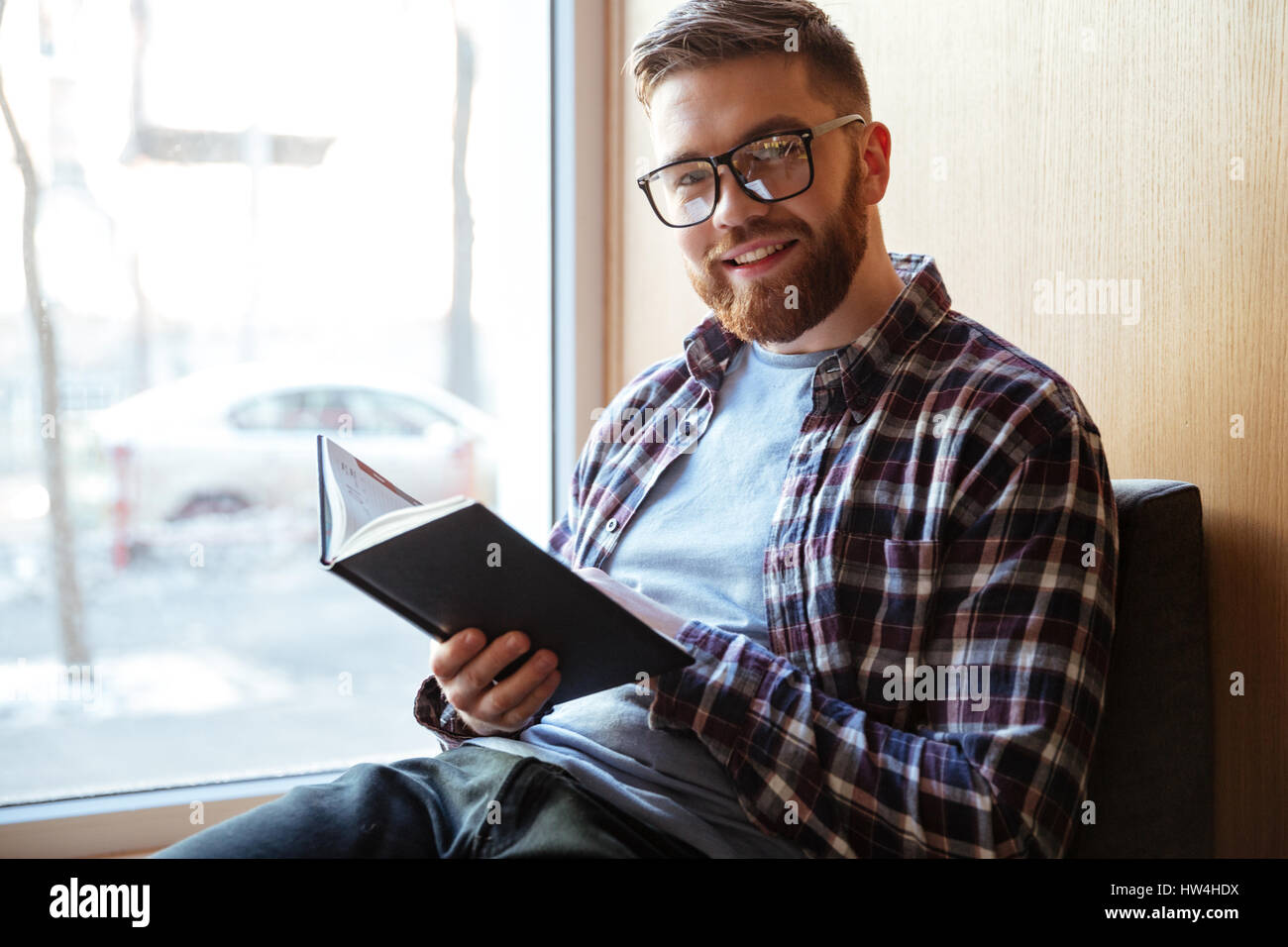 Porträt eines lächelnden glücklich männlichen Studenten Buch hält und Blick in die Kamera beim Sitzen auf der Fensterbank in Bibliothek Stockfoto