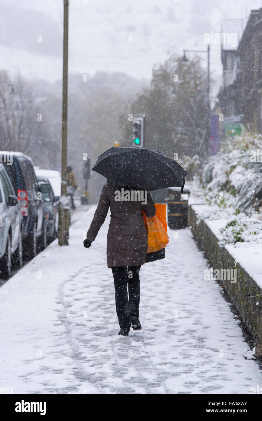 Es schneit an einem kalten Wintertag und eine Dame (trägt einen langen Mantel und hält einen Regenschirm) Spaziergänge auf dem Bürgersteig - Ilkley, West Yorkshire, GB. Stockfoto