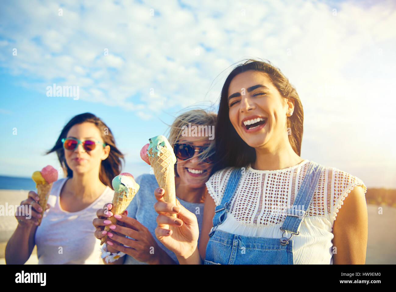 Glücklich temperamentvolle junge Studentinnen zu Fuß entlang einer Strandpromenade essen Eiscreme-Kegel, lachen und scherzen in der heißen Sommersonne Stockfoto
