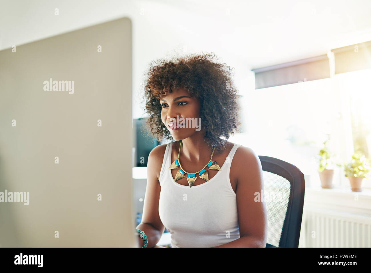 Lächelnde junge afrikanische Unternehmerin sitzt an ihrem Schreibtisch in ihrem Büro zu Hause arbeiten auf einem großen Desktop-Monitor, hoher wichtigen Hintergrund Stockfoto