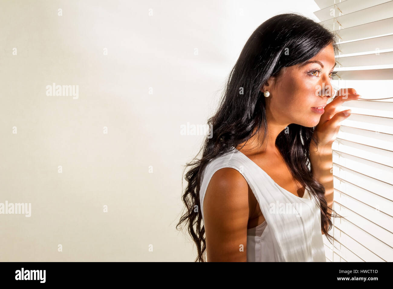 Eine junge Frau beobachtet etwas durch die Jalousie von ihrem Fenster, Eine Junge Frau Beobachtet Etwas Durch Die Jalousie Ihres Fensters Stockfoto