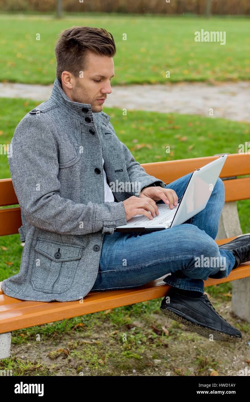 Ein Mann mit einem Laptop sitzt auf einer Parkbank, Ein Mann Mit Einem Laptop Sitzt Auf Einer Parkbank Stockfoto