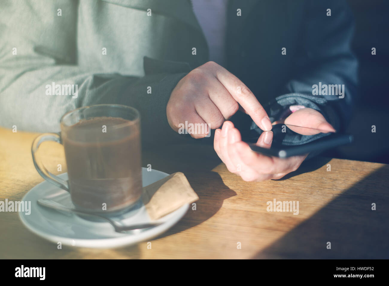 Frau mit Handy und trinken heißen Schokolade im Café. Lässige jungen kaukasischen Erwachsenfrau hinter dem Glas Textmitteilung zu schreiben, auf Smartphone ap Stockfoto