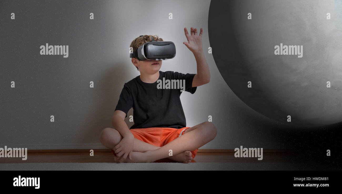 Kleiner Junge Schneidersitz sitzen, virtual-Reality-Kopfhörer tragen, ausstrecken, Planet, digital Composite zu berühren Stockfoto