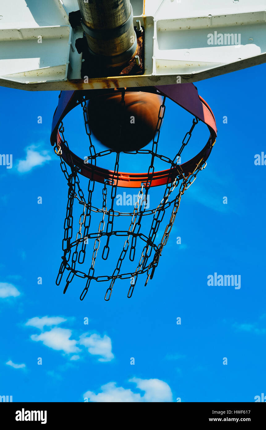 Einen Basketballkorb mit einem Basketball. Die Basketball geht in den Reifen. Der Reifen hat ein doppelter Rand und einer Kette netto. Es wird von unten mit dem Himmel geschossen. Stockfoto