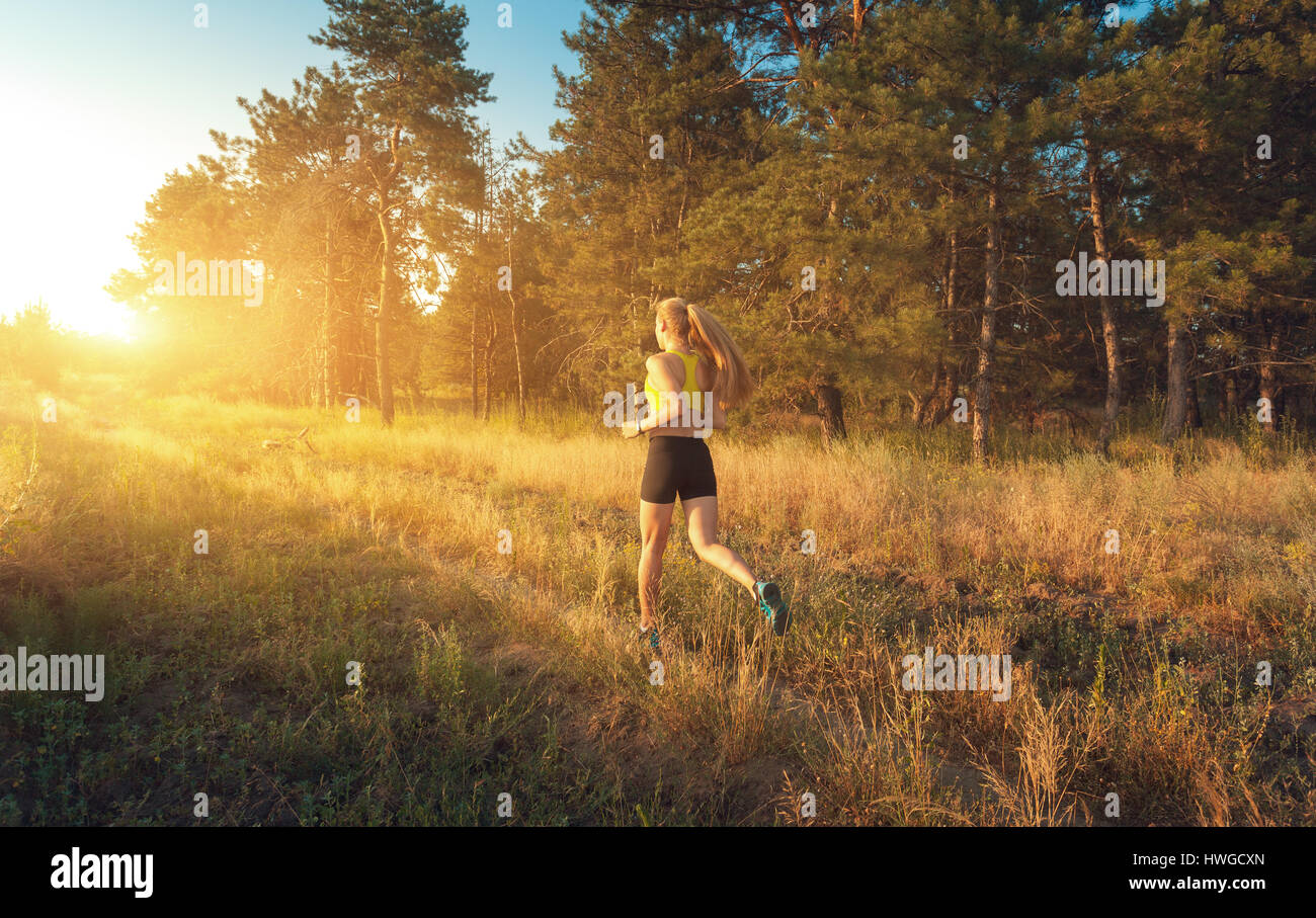 Sportliche Mädchen läuft auf einem Feld nahe der Bäume bei Sonnenuntergang im Sommer. Athlet läuft auf der off-Road am Abend. Aktive Frau. Sport und Gesundheit Stockfoto