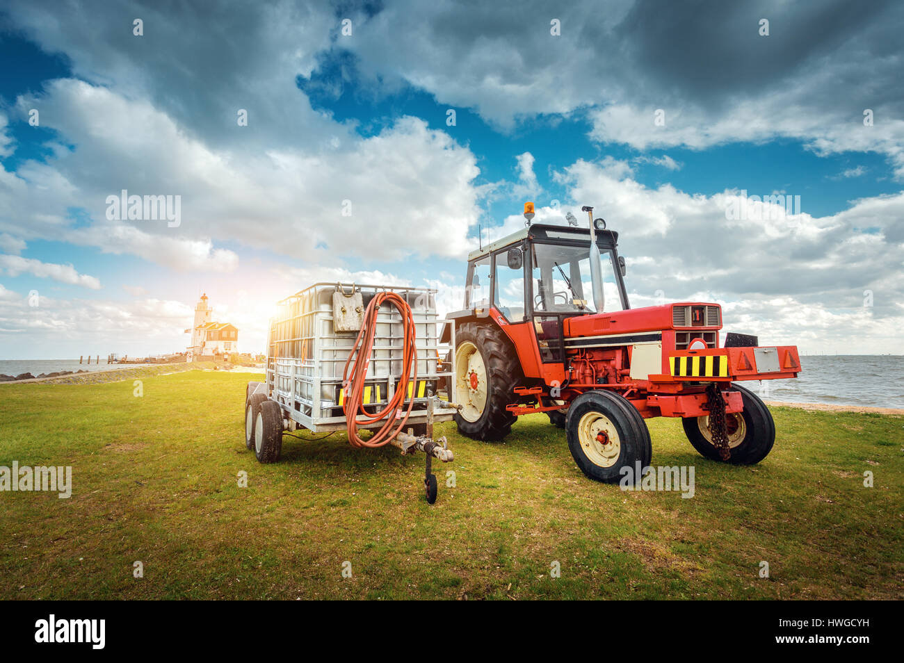 Roter Traktor mit Anhänger auf der Wiese gegen Leuchtturm und blau bewölktem Himmel im Frühjahr bei Sonnenuntergang. Landwirtschaftliche Zugmaschine. Landwirtschaftliche Maschinen ein Stockfoto