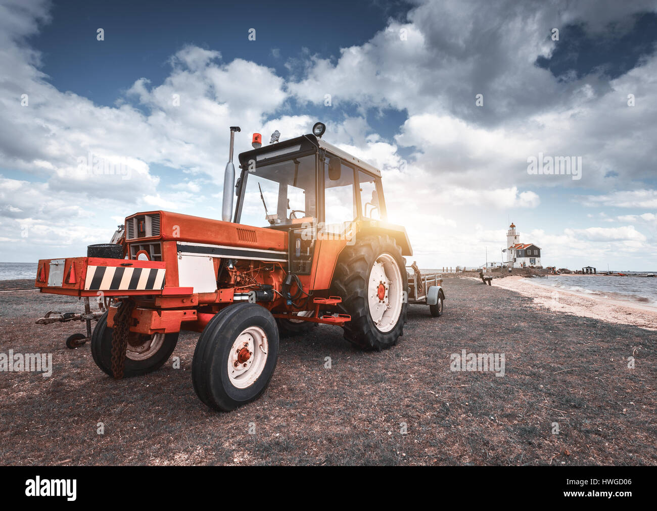 Roter Traktor auf der Wiese gegen Leuchtturm und blau bewölktem Himmel im Frühjahr bei Sonnenuntergang. Landwirtschaftliche Zugmaschine. Landwirtschaftliche Maschinen und Maschinen ich Stockfoto