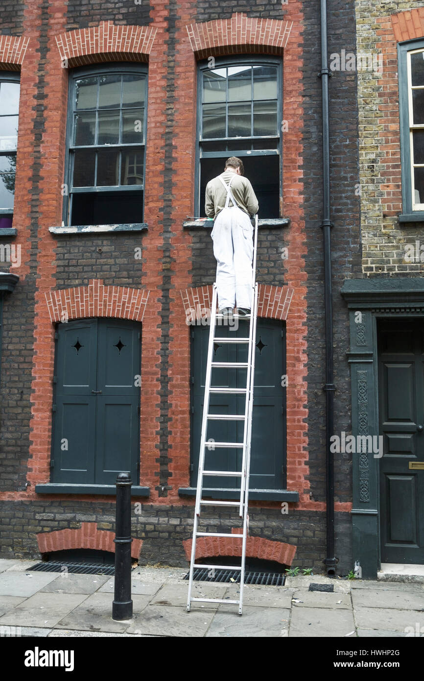 Der Handwerker gibt den Mann an einer Leiter ein, die den Fensterrahmen eines Londoner Huguenot-Hauses im Osten Londons, Großbritannien, repariert. Selbstständige Erwerbstätige in Großbritannien. Händler Großbritannien. Fenster „Leiter“. Stockfoto