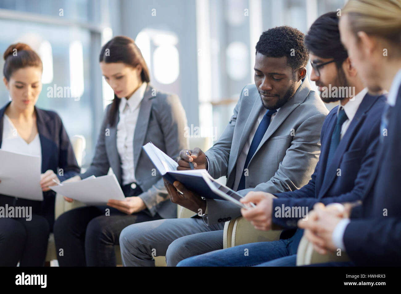 Multi-ethnischen Gruppe von Geschäftsleuten, die sitzen in Reihe in modernen Glashalle, Fokus auf afrikanisch-amerikanischer Geschäftsmann Handout Lesematerial und machen Stockfoto