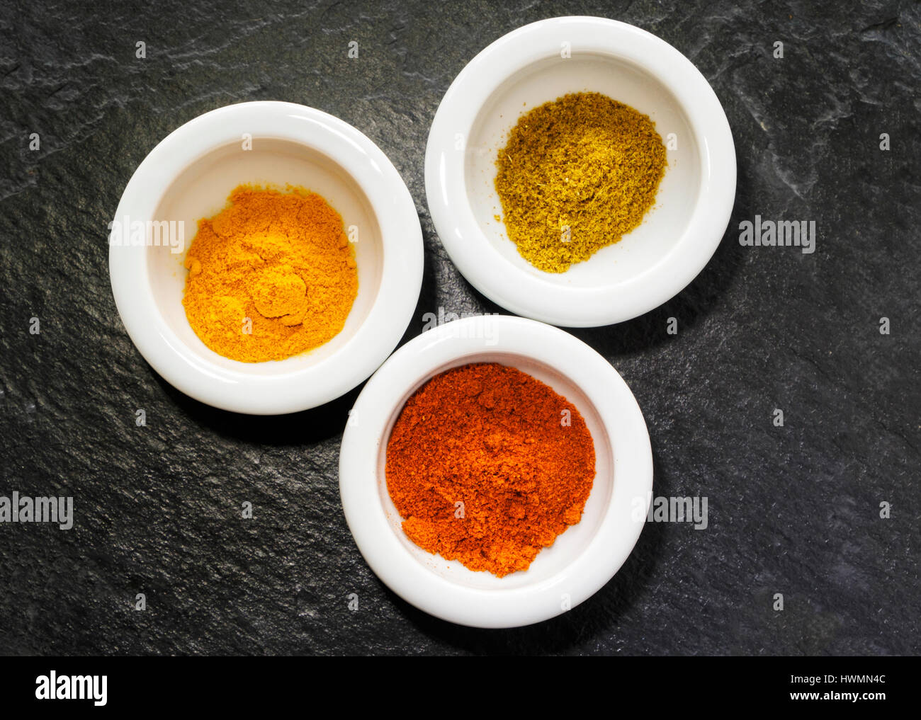 Lay-flat-Bild der drei kleine weiße Geschirr mit bunten Gewürzen auf strukturiertem schwarzem Schiefer. Kurkuma, Kreuzkümmel und Paprika. Stockfoto