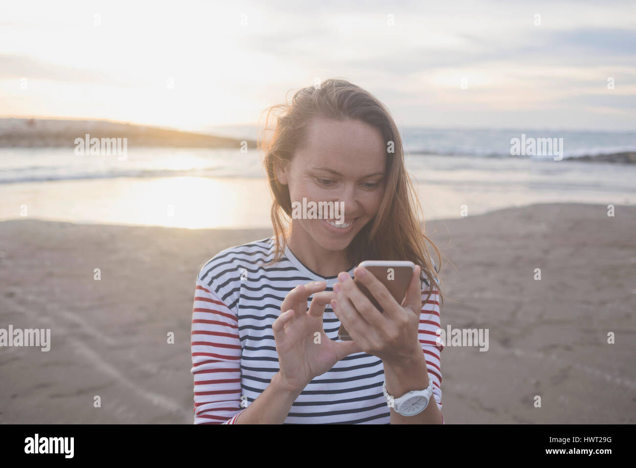 Lächelnde Frau mit Handy am Strand bei Sonnenuntergang Stockfoto