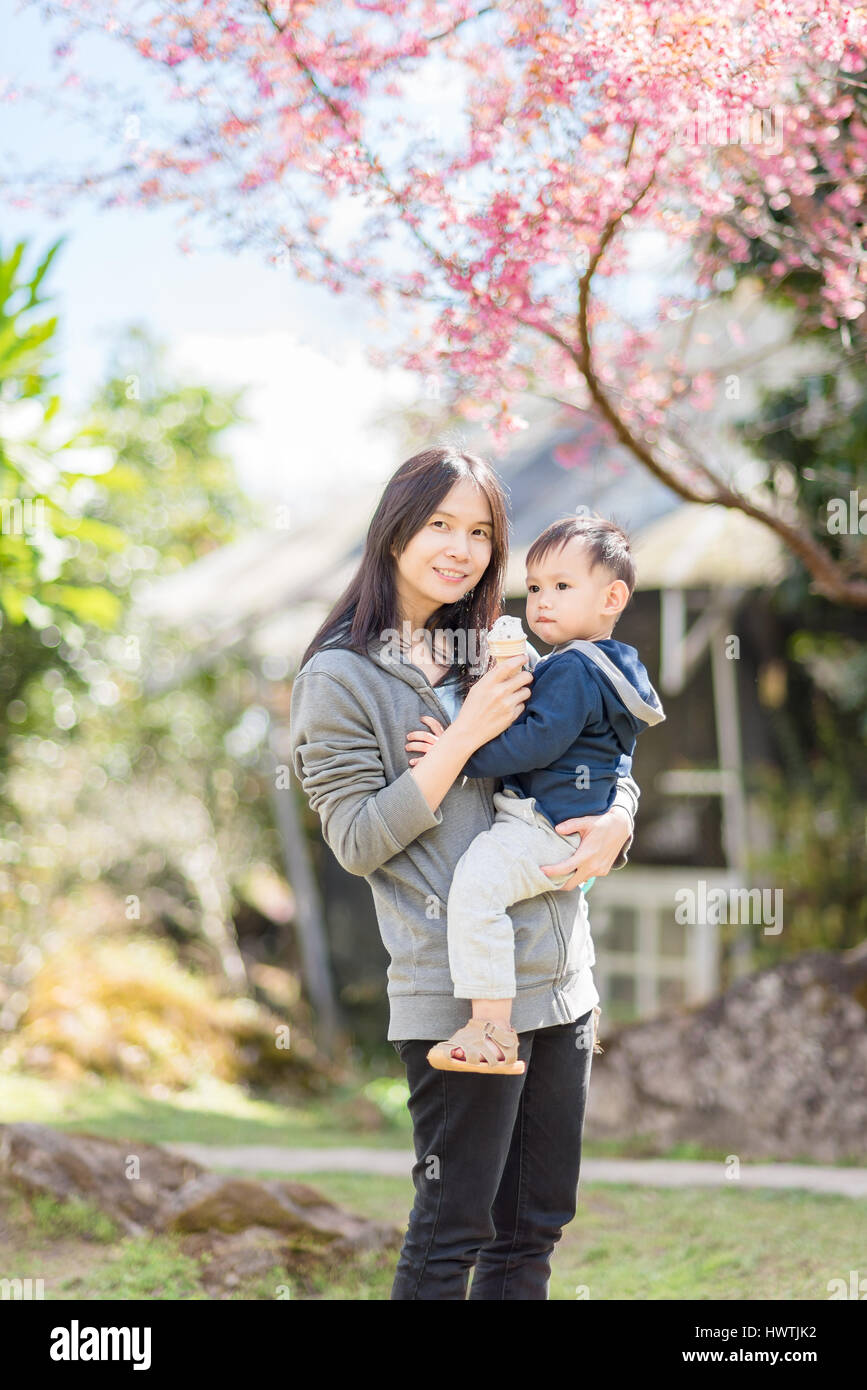 Asiatische Mutter halten Baby im Garten blühen Kirschblüten. Mutter und Sohn gemeinsam essen Eiscreme-Kegel Stockfoto