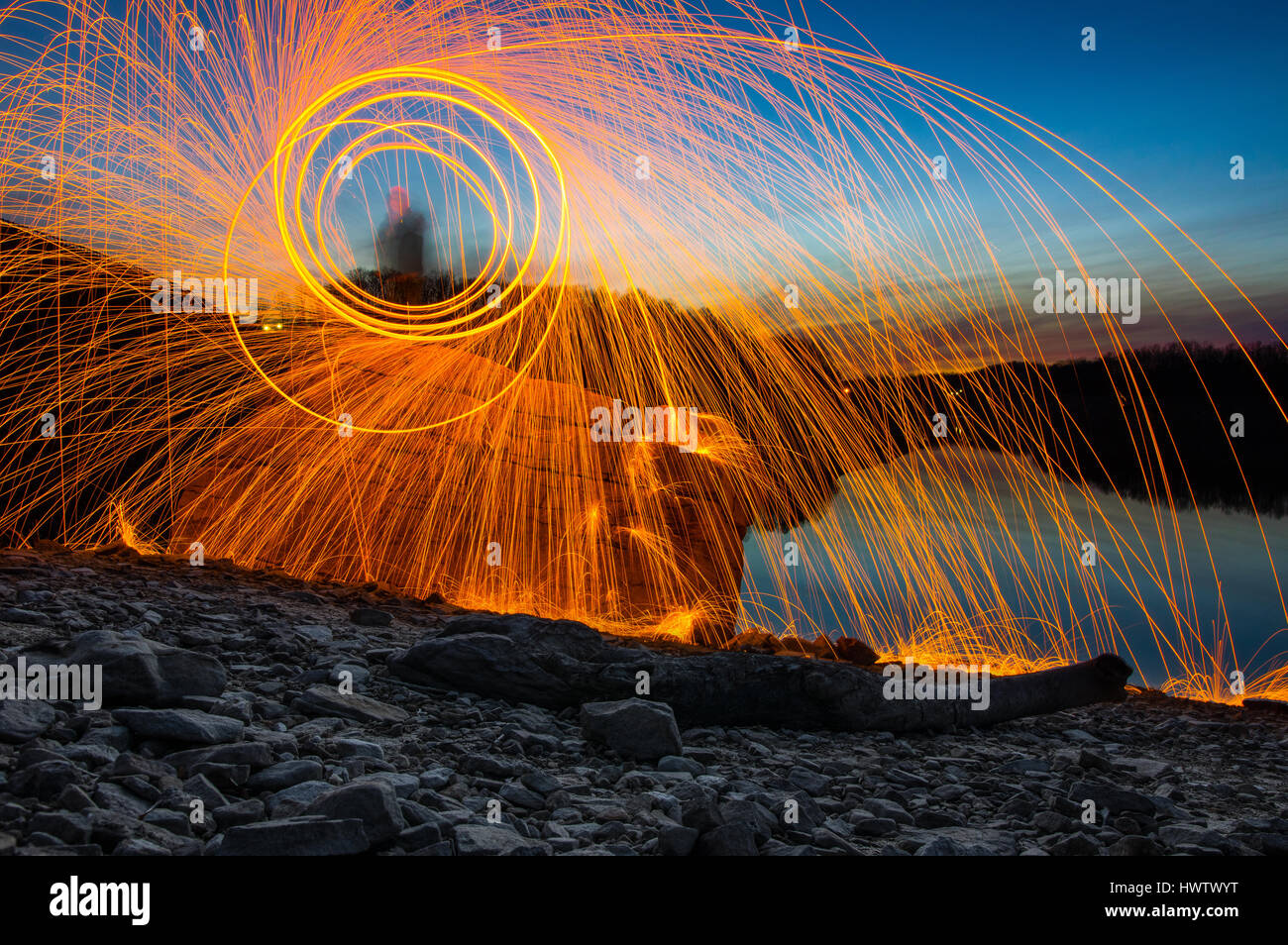 Eine Person steht auf einem Felsen gegen einen See in der Abenddämmerung Spinnerei Stahlwolle auf Feuer mit fliegen in alle Richtungen Funken. Stockfoto
