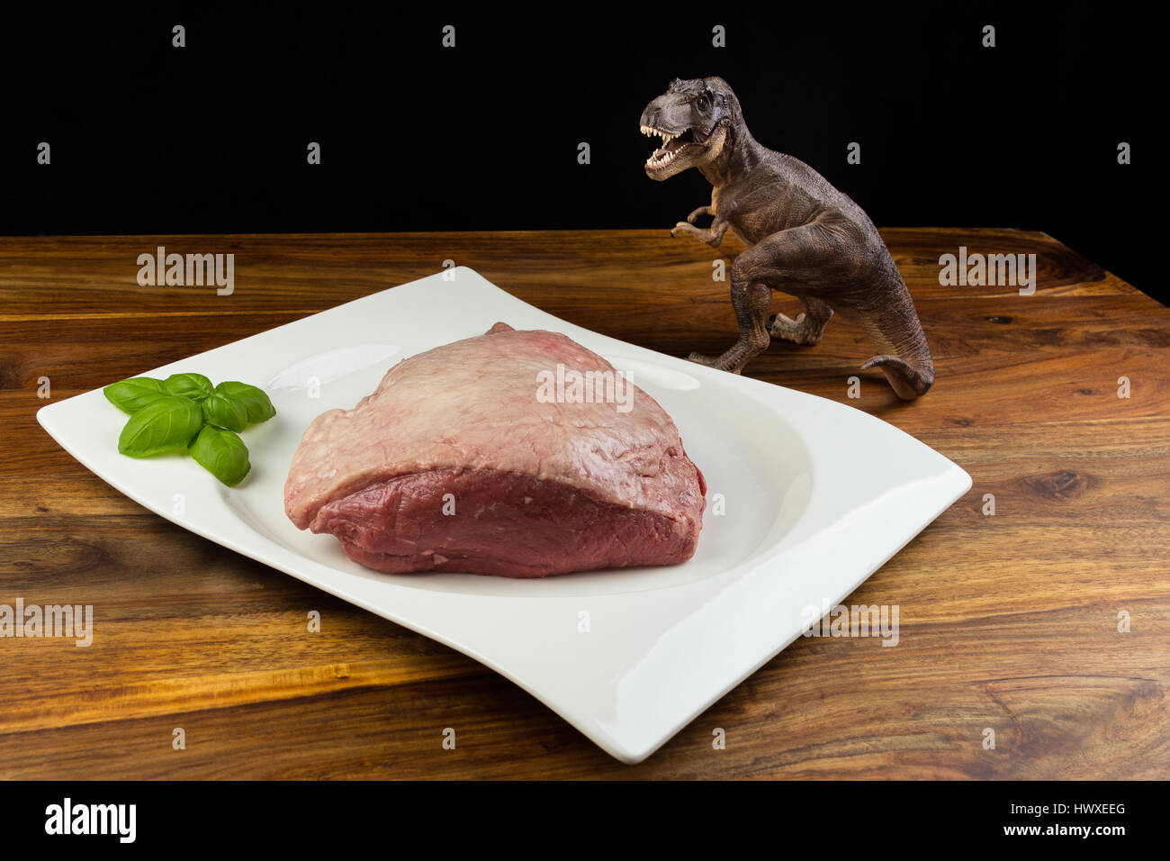 Dinosaurier Tyrannosaurus Rex starrte ein komplettes Stück roh Kuh Rindfleisch (Picanha), mit Basilikum auf einem weißen Teller und natürliche Holztisch - dunklen Hintergrund Stockfoto