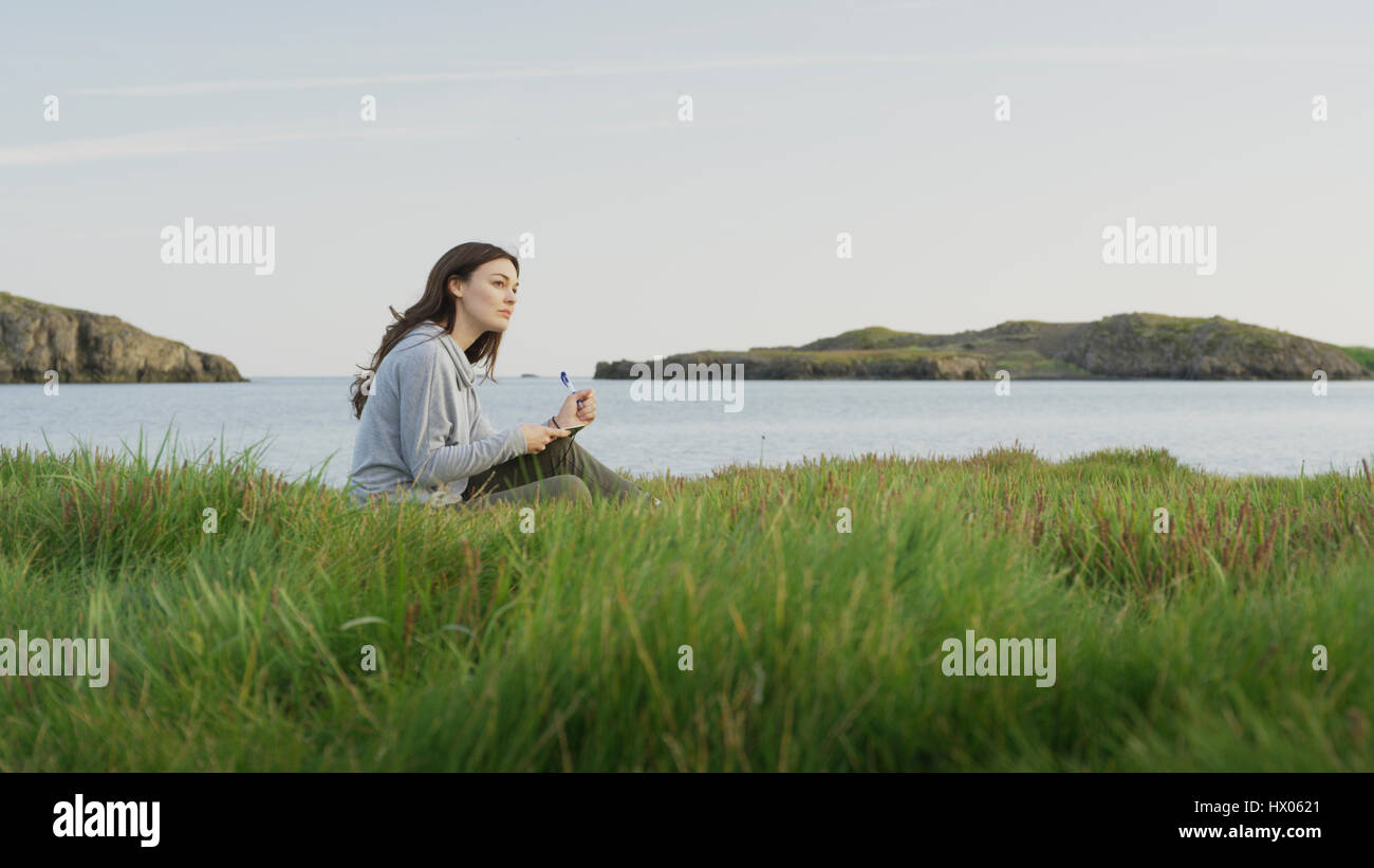Profil von Frau sitzt auf Wiese in der Nähe von See in abgelegenen Landschaft unter blauem Himmel Stockfoto