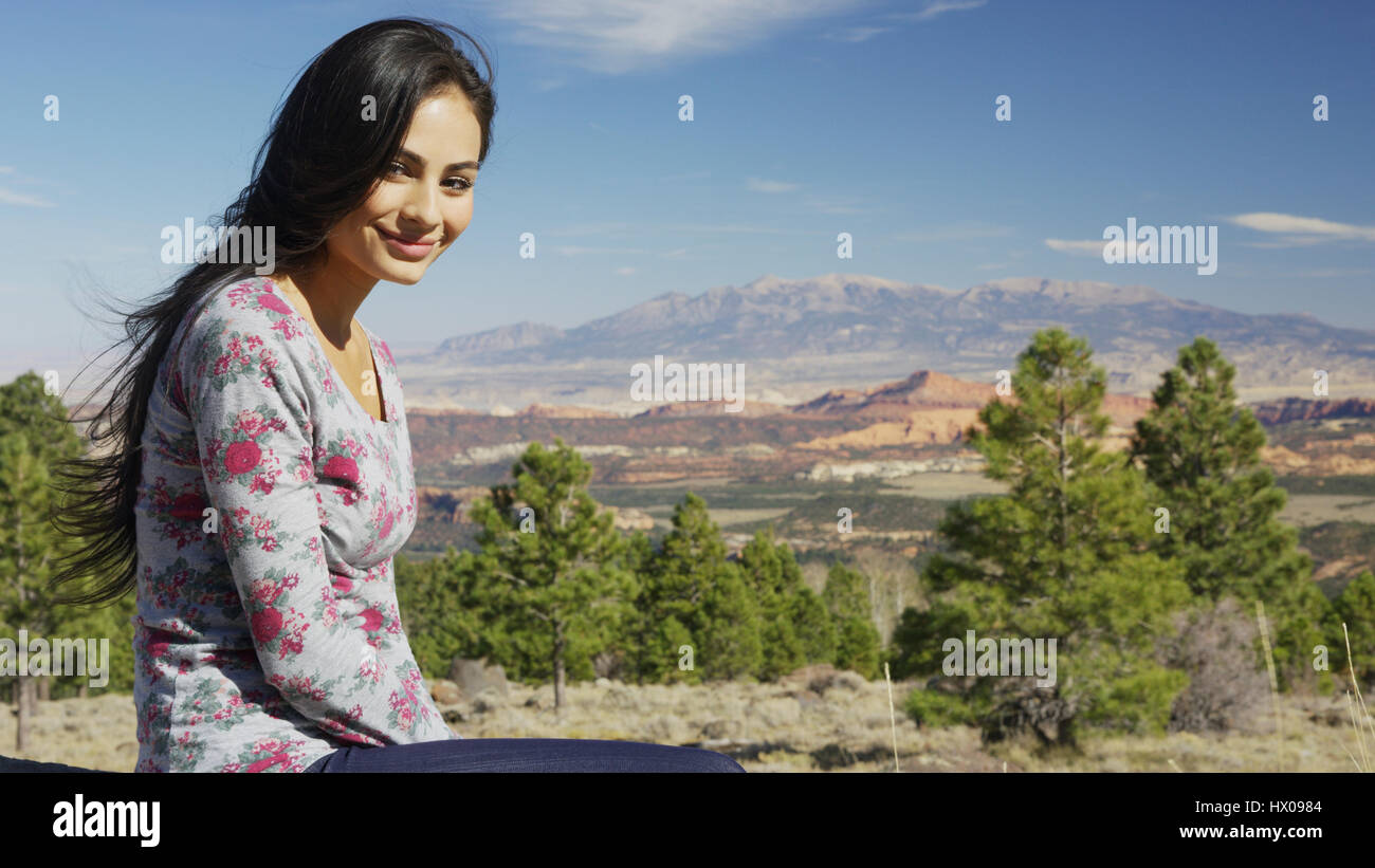 Porträt von lächelnden Frau sitzt in der Nähe von malerischen Blick auf Berge, Bäume und Buschwerk in abgelegenen Landschaft unter blauem Himmel Stockfoto