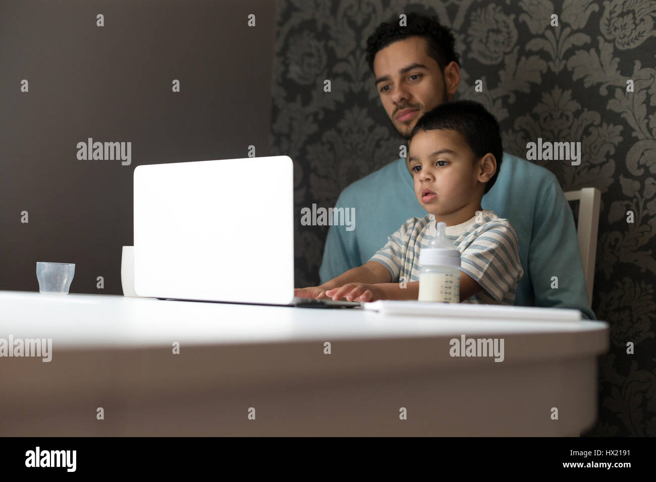 Junger Vater sitzt am Tisch mit seinem Sohn auf dem Schoß. Sein Sohn ist kopieren sein Vater und seine Neugier zu erkunden, durch Drücken der Tasten und auf der Suche nach einem Stockfoto