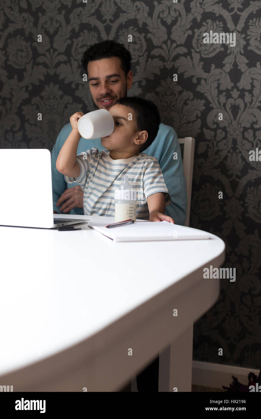 Junger Vater sitzt am Tisch mit seinem Sohn auf dem Schoß. Sein Sohn ist kopieren, was er gesehen hat, seinen Vater zu tun und hat seine Väter Becher an den Mund, Stockfoto