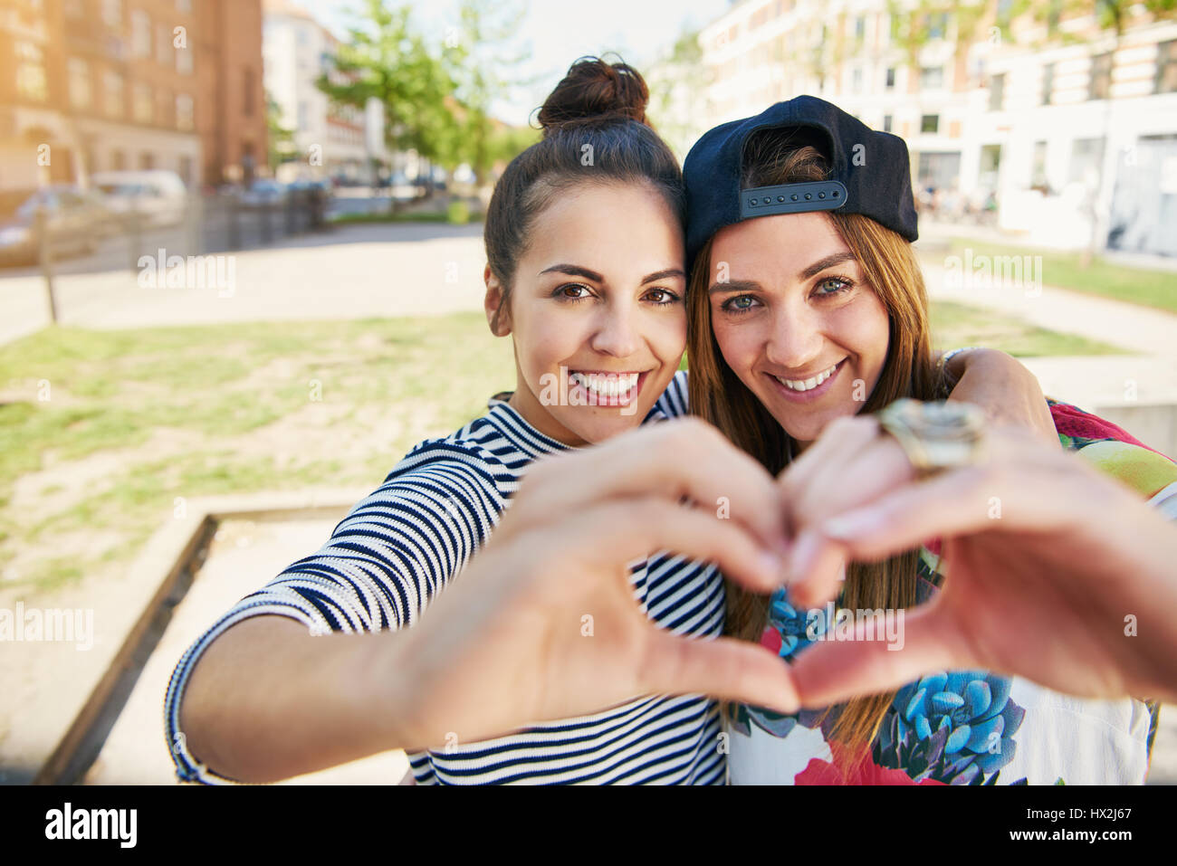 Zwei romantische junge Freundinnen ein Herz Geste kombinieren ihre Hände als die Pose arm in arm in die Kamera grinsend Stockfoto