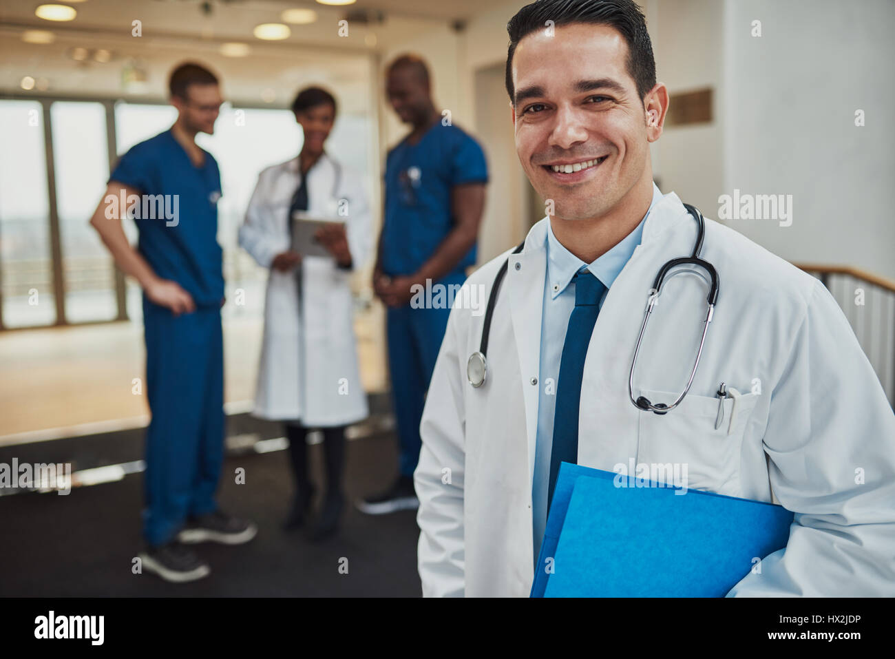 Erfolgreiche hübscher junger Hispanic Arzt mit einem strahlenden Lächeln stehen umklammert einen blauen Ordner unter dem Arm mit seinem Ärzteteam im Hintergrund Stockfoto