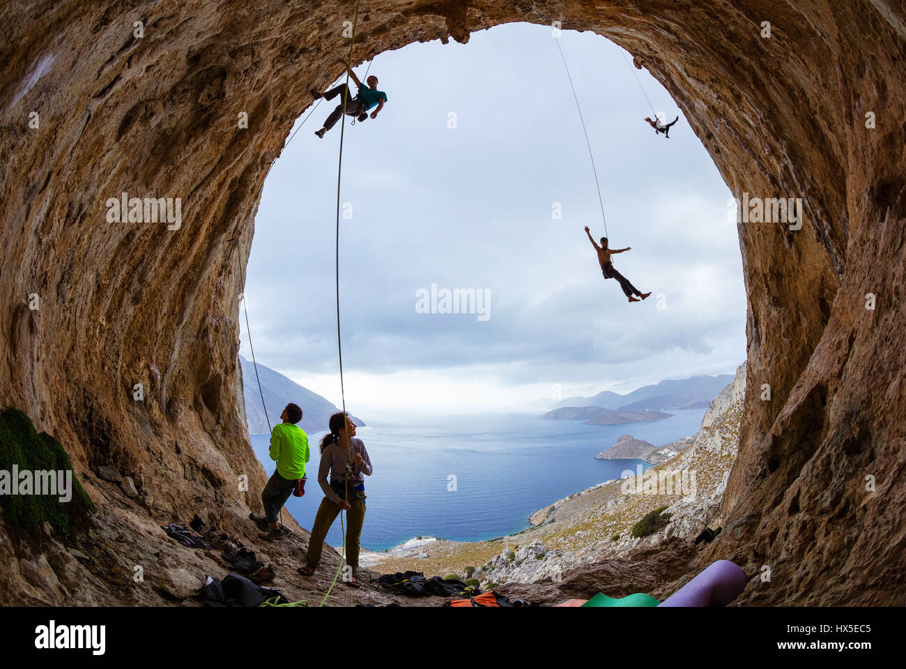 Sportkletterer in Höhle: Belayers führenden Bergsteiger, zwei Kletterer schwingen auf Seilen oder abgesenkt werden, nach unten, eine männliche packende Haltegriff auf Felsen beobachten ein Stockfoto