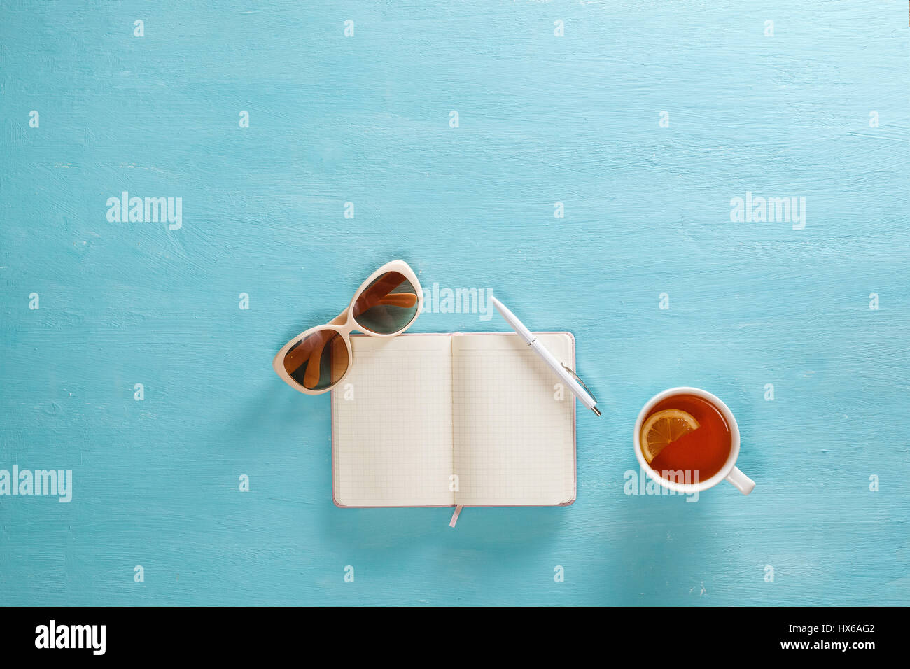 Notizbuch mit Stift, Tasse Tee und Sonnenbrille auf blauen Holztisch eröffnet. Ansicht von oben. Schreiben, Bloggen Konzept. Kopieren Sie Raum. Stockfoto