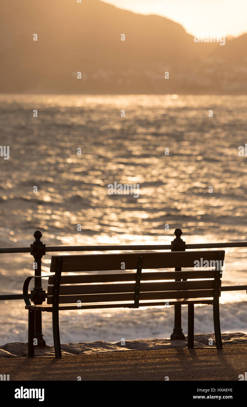 Holzbank an der Strandpromenade von den beliebten Ferienort Stadt Llandudno in Nordwales wie die Sonne beginnt zu steigen über den little Orme entlang der Küste Stockfoto